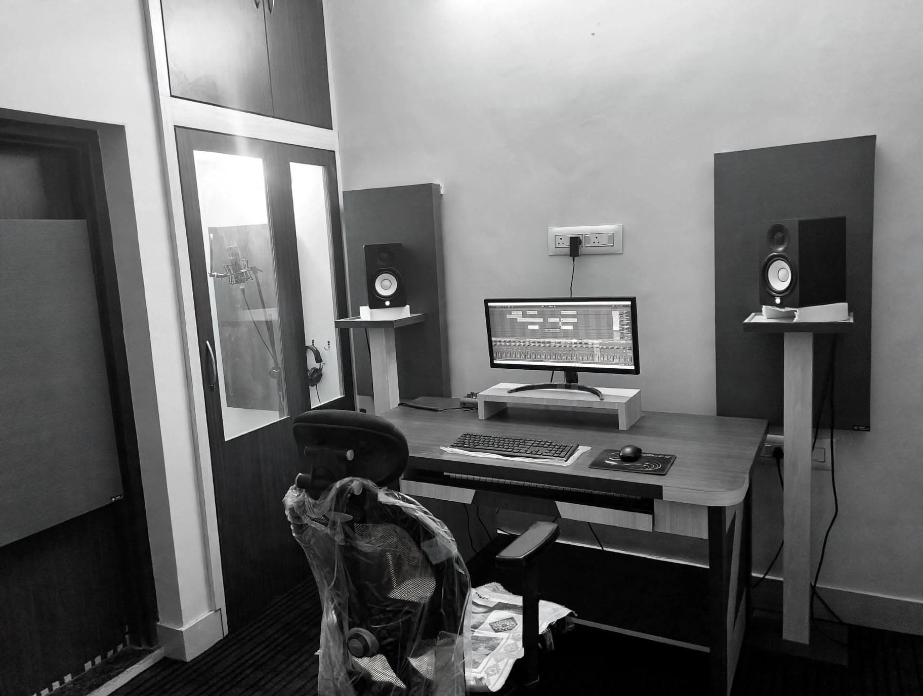 Studio desk and monitor with the recording booth and microphone visible in the background
